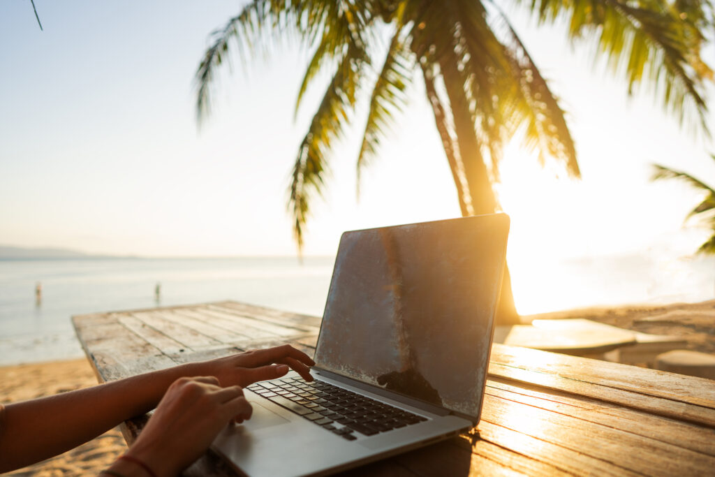 An image of a girl freelancer sitting on a rock by the sea at sunset, typing on her laptop. This image represents five types of remote workers: a freelancer or independent contractor, who works for themselves and can work from anywhere; a virtual employee, who can work from home or any remote location with stable pay and benefits; a content creator, who can use their creativity to make digital content for an online audience; a virtual coach or consultant, who can work with clients from anywhere to help them reach their goals; and a virtual entrepreneur, who can turn an idea into a thriving online business while living life on their own terms.